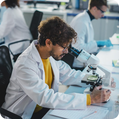 A man seated at a table while using a microscope. Other people are seated at other tables in the background